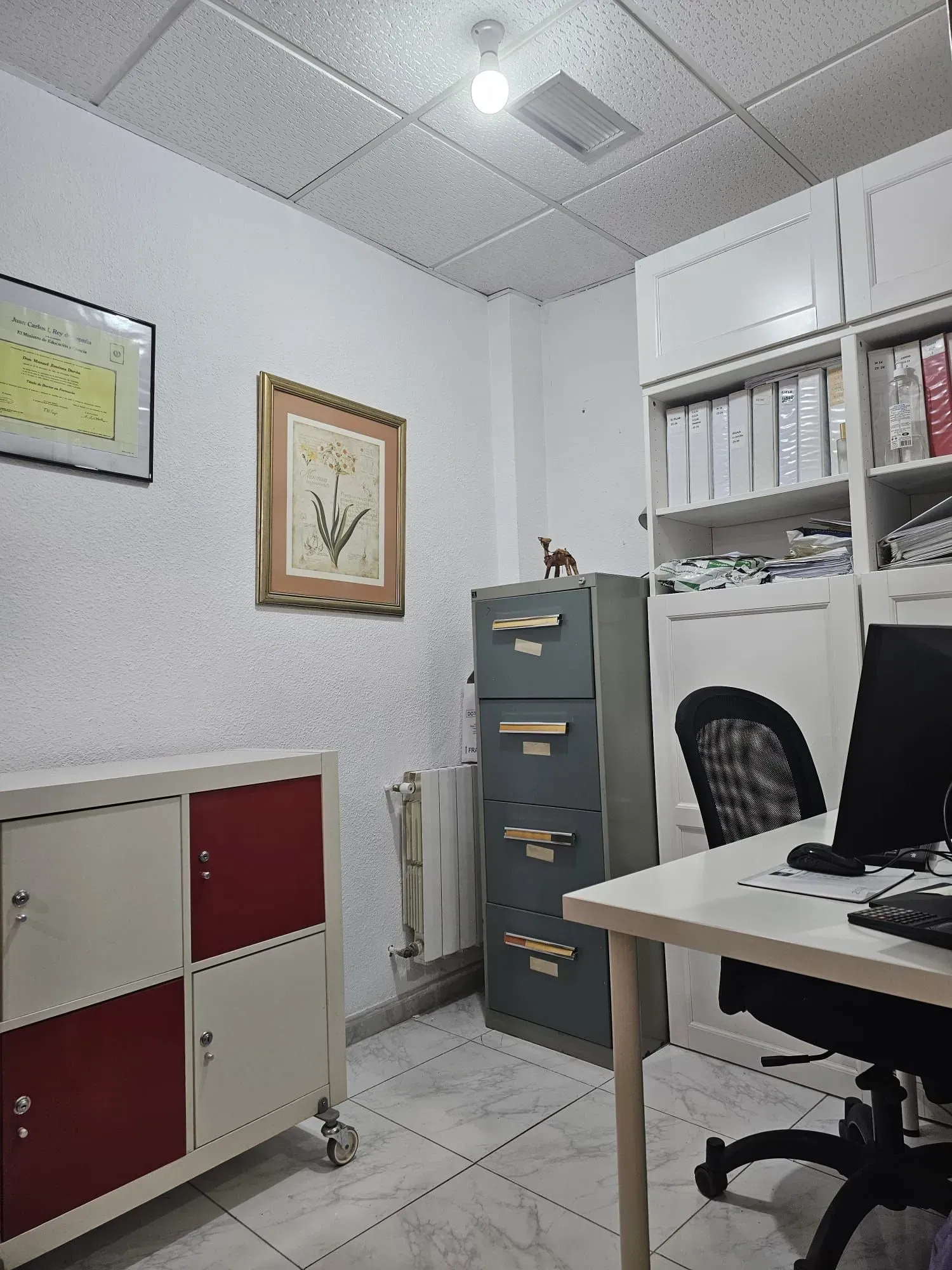 Oficina con escritorio, archivador, armarios blancos, arte enmarcado y una silla.