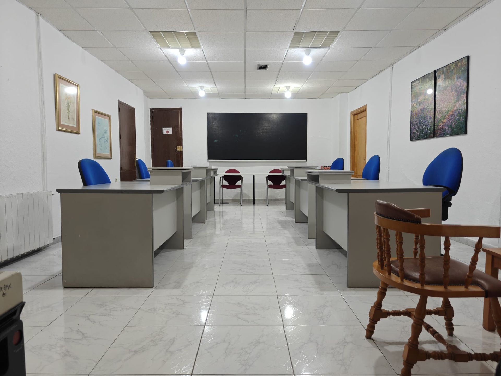 Aula vacía con mesas, sillas y pizarra. Paredes blancas, suelo de baldosas y dispensador de agua.