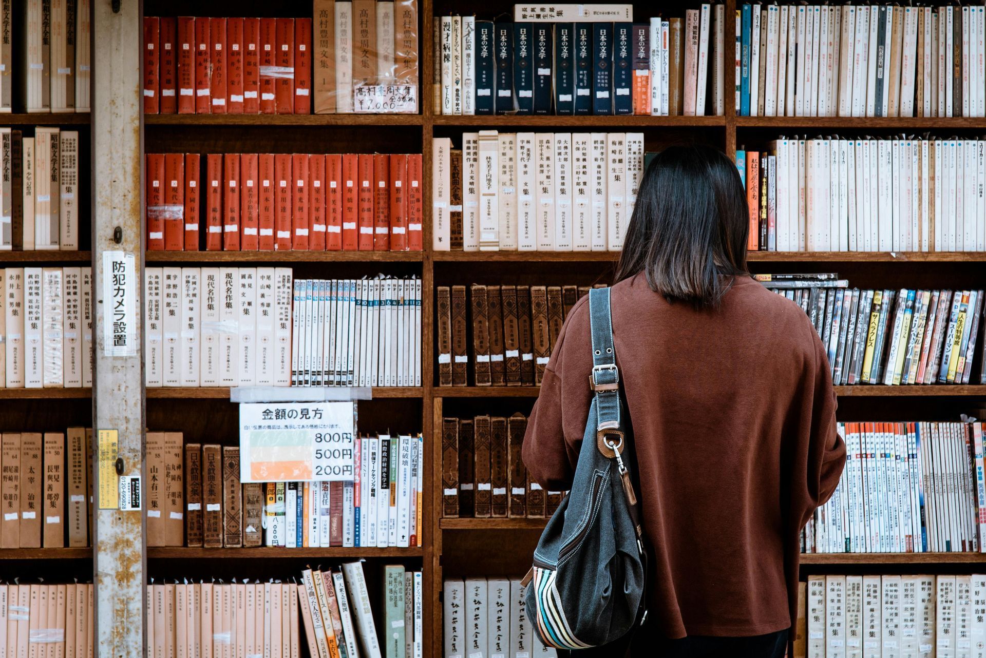 Persona hojeando estanterías de libros en una biblioteca, vistiendo un suéter marrón y un bolso de hombro.