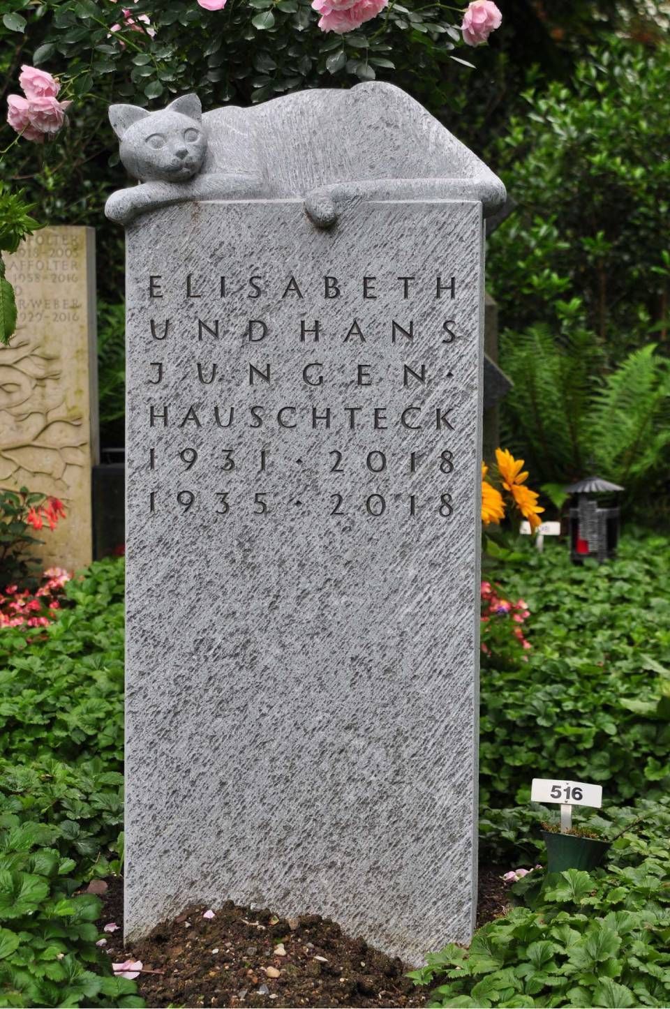 Grauer Granitgrabstein in einem Garten mit einer Katzenskulptur auf der Spitze, umgeben von Blumen.