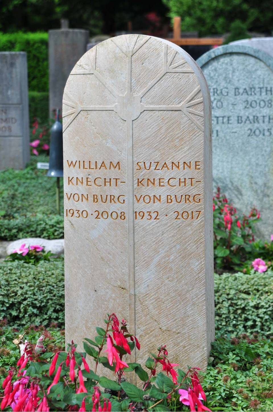 Zwei gravierte Grabsteine ​​mit Blumen davor, umgeben von Grün.