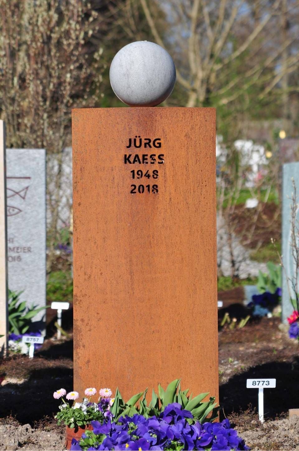 Rostfarbener Grabstein mit einer weissen Kugel an der Spitze, eingraviert „Jörg Kaese 1948 2018“, umgeben von Blumen.