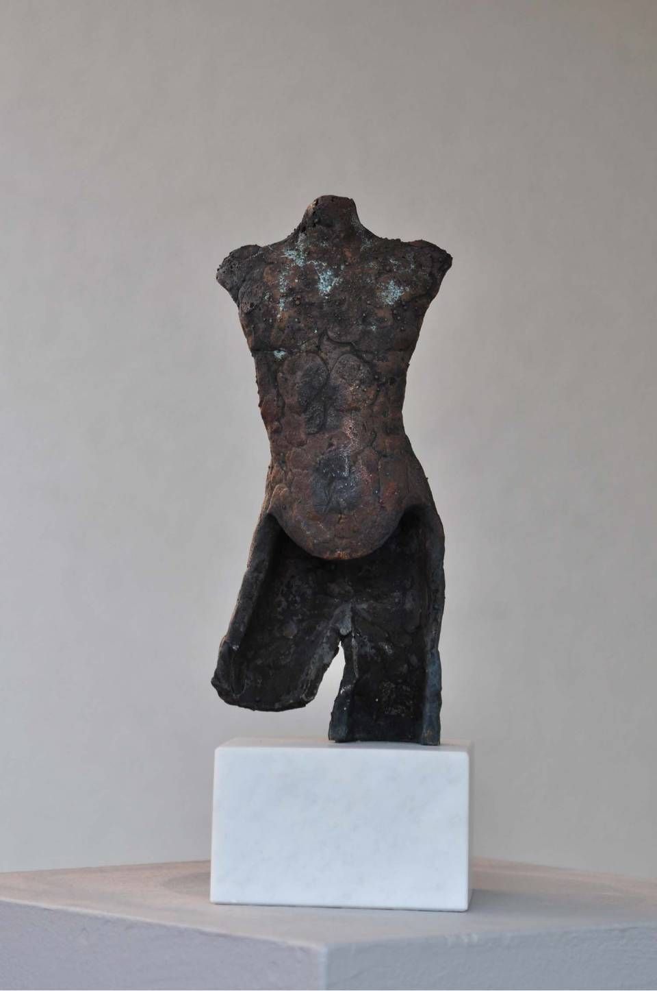 Kleine abstrakte Skulptur aus dunklem Metall auf einem weissen Sockel vor einer schlichten Wand.