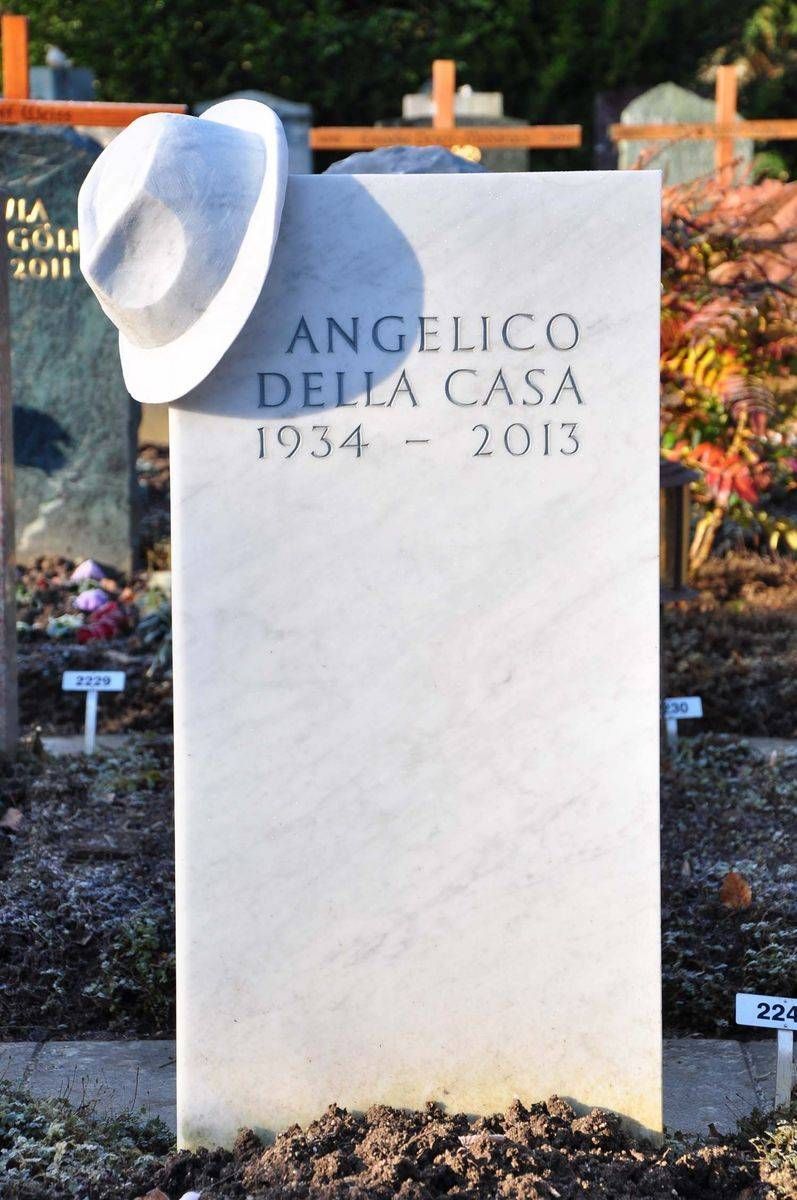 Weisser Grabstein mit Hut darauf, eingraviert „Angelo Della Casa 1914–2013“, auf einem Friedhof.