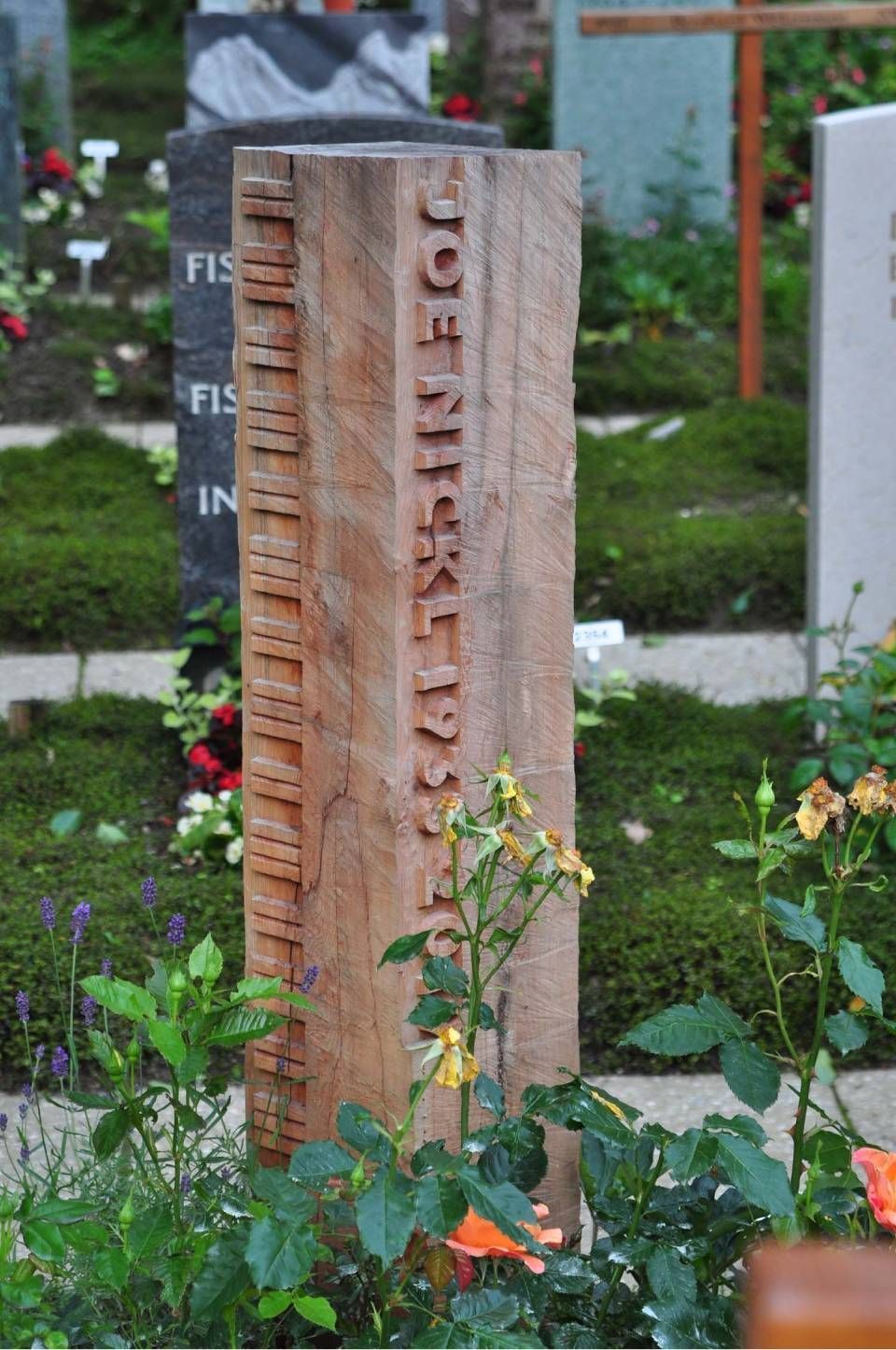 Vertikales Steinmonument in einem Garten, mit Inschrift versehen und von Blumen und Grün umgeben