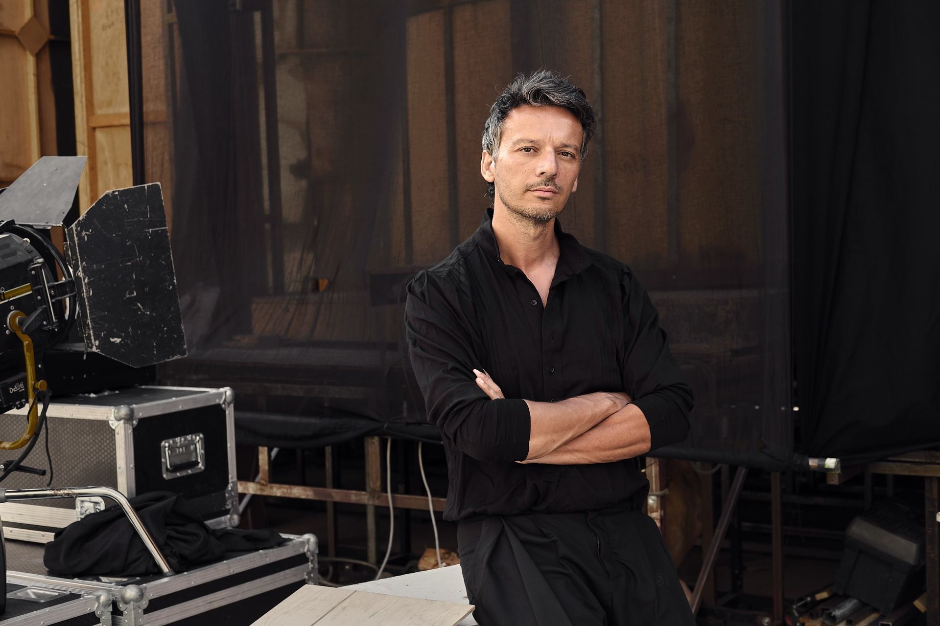 Άνδρας με σταυρωμένα χέρια, φορώντας μαύρα ρούχα, στέκεται σε ένα στούντιο με φωτιστικό εξοπλισμό και μαύρο φόντο.