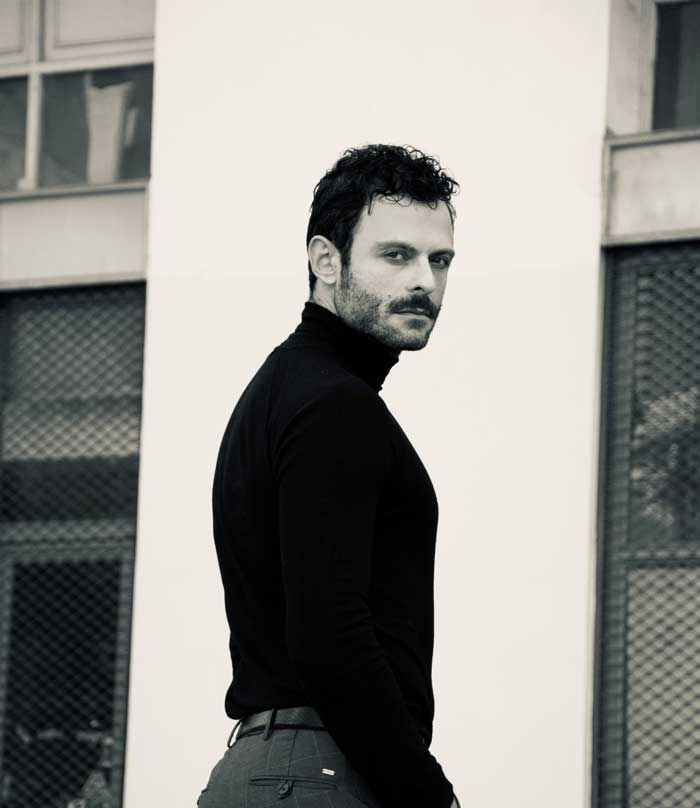 Άντρας με μαύρο ζιβάγκο κοιτάζει πάνω από τον ώμο, στέκεται κοντά σε ένα κτίριο.