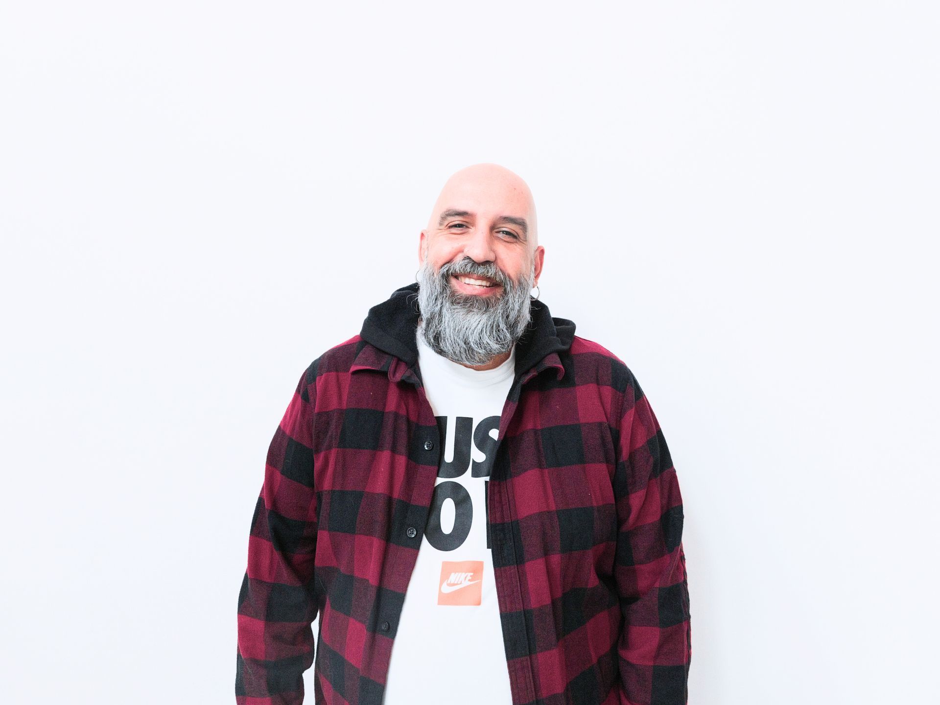 Φαλακρός άντρας με γκρίζα γενειάδα που φοράει κόκκινη και μαύρη φανέλα πάνω από ένα λευκό μπλουζάκι Nike, χαμογελάει.