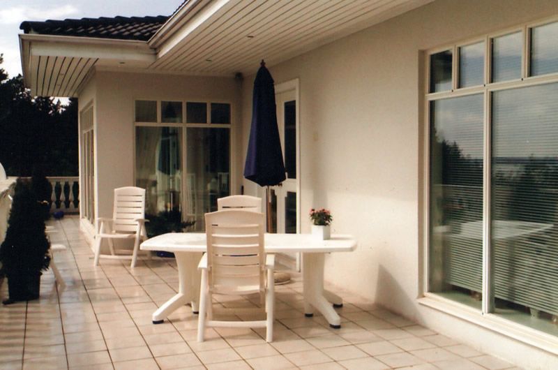 Patio, jolla on valkoinen pöytä ja tuolit talon vieressä, tumma aurinkovarjo ja suuret ikkunat.