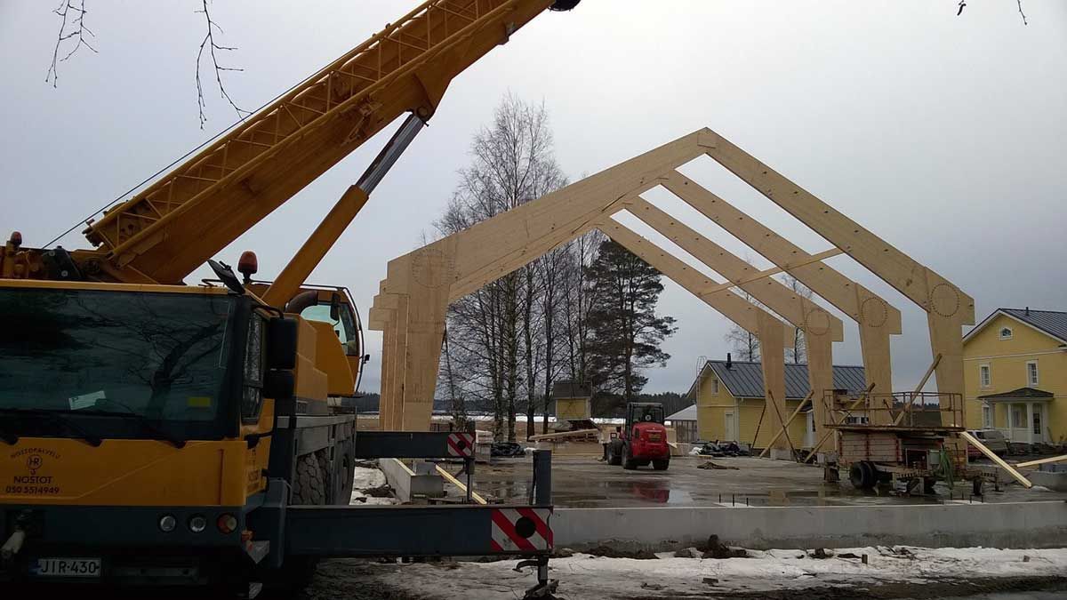 Rakennustyömaa, jossa on keltainen nosturi ja suuret puiset holvikaarikehykset lumisessa maisemassa