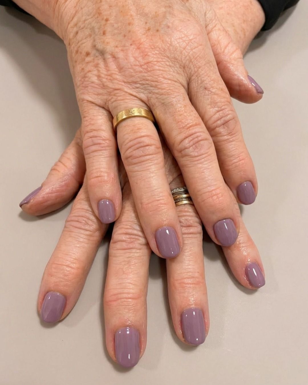 Manos con esmalte de uñas malva y anillos de oro descansando juntas sobre una superficie clara.