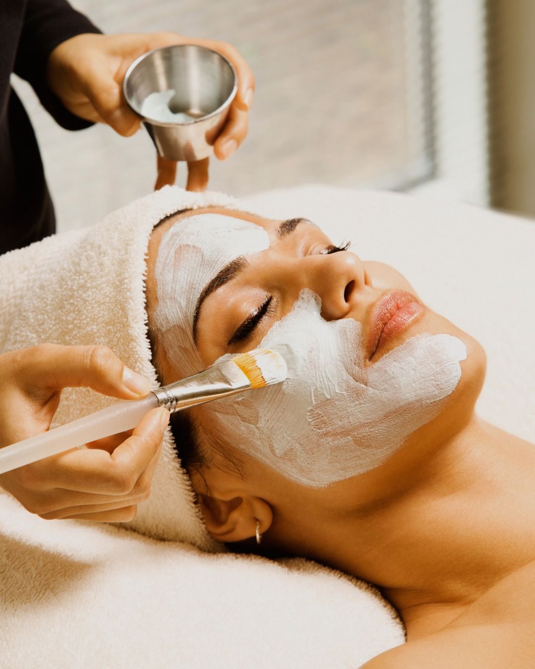 Tratamiento facial de spa con aplicación de crema blanca mediante brocha sobre el rostro de una persona relajada.
