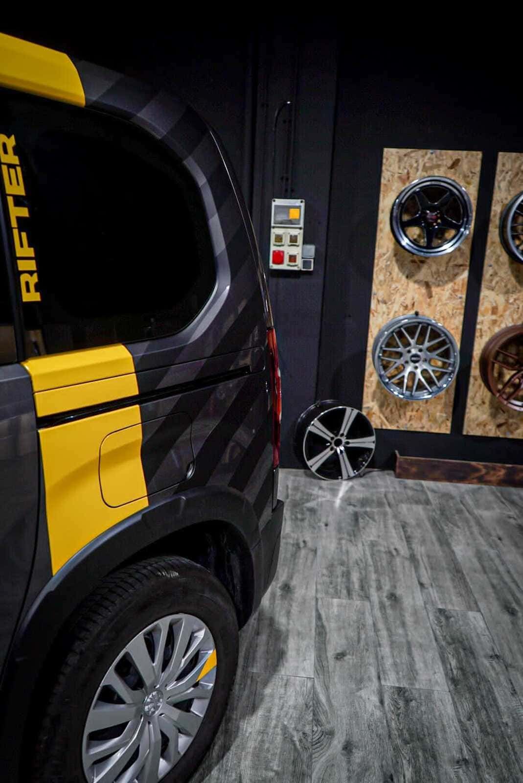 Una furgoneta negra y amarilla está aparcada en un garaje junto a una pared de ruedas.