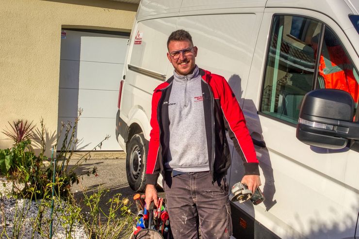 Un homme, vêtu d'une veste rouge et tenant des outils, se tenait à côté d'une camionnette blanche, à l'extérieur, près d'une maison.
