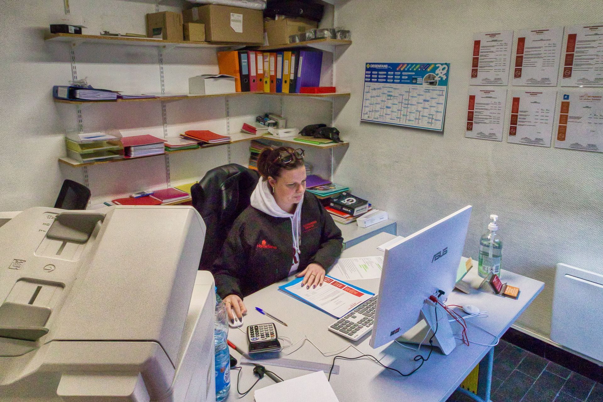 Une personne travaille à un bureau dans un bureau encombré, utilisant un ordinateur à côté d'une imprimante et d'étagères murales.