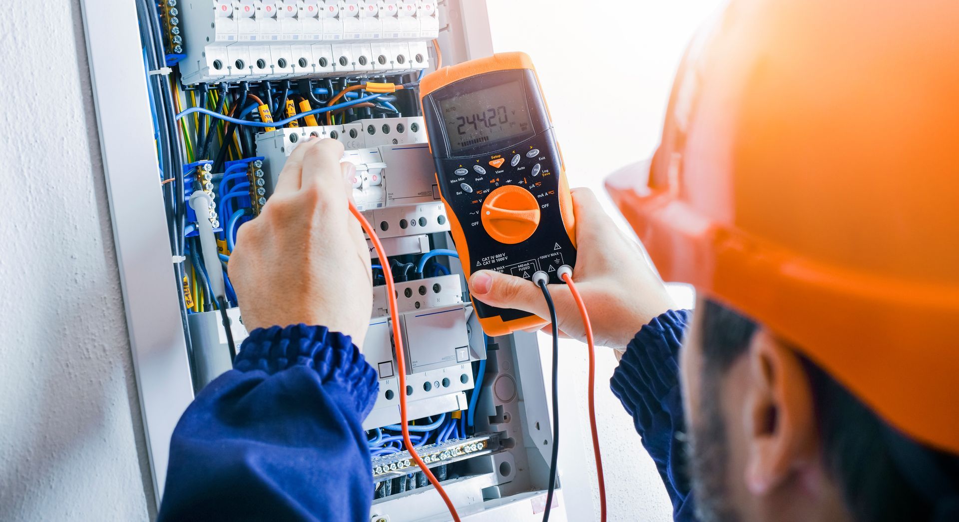 Technicien portant un casque orange testant un tableau électrique avec un multimètre.