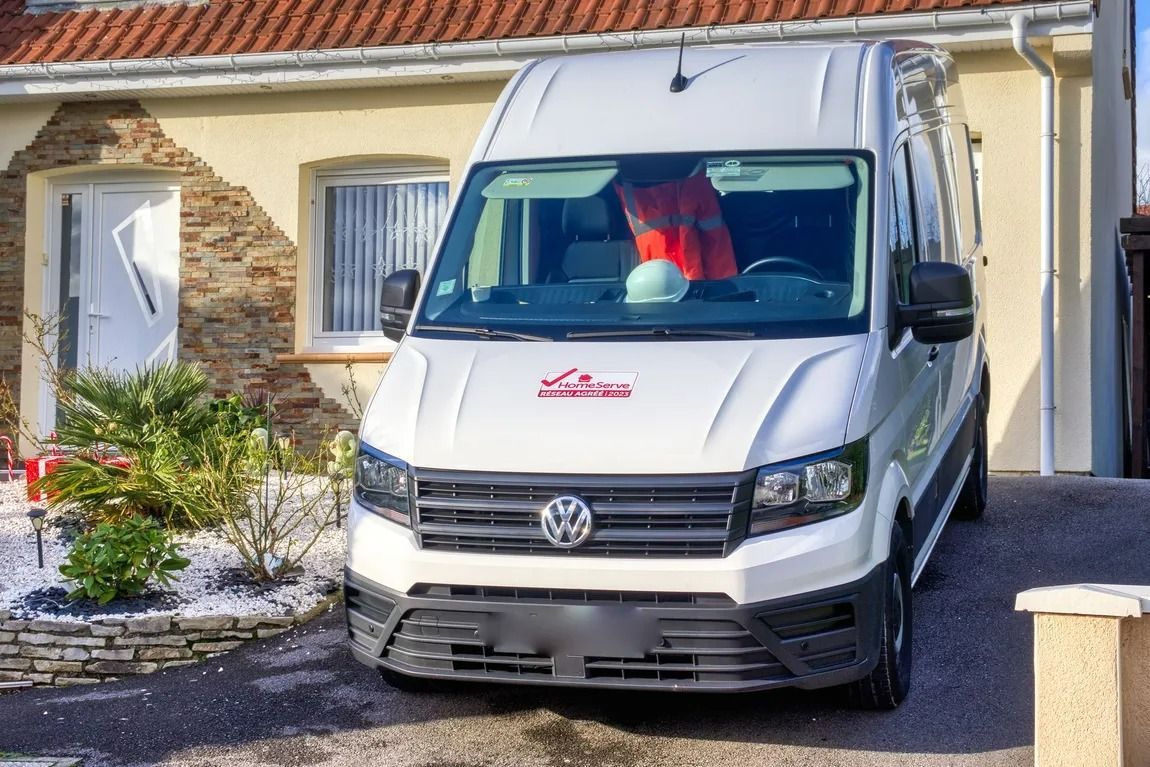Camionnette de livraison blanche garée devant une maison au toit de tuiles rouges et aux murs en pierre.