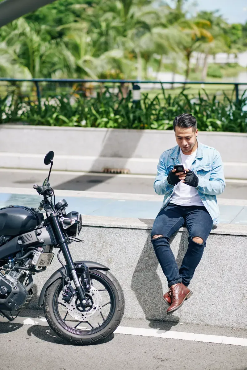 Hombre con motocicleta usando un teléfono, sentado en una pared en un entorno soleado al aire libre.