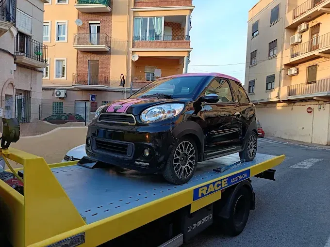 Automóvil blanco con daños en la parte delantera en una calle de la ciudad; guardabarros dañado, neumático visible.
