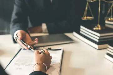 Persona firmando documento en una mesa con un abogado, balanza de la justicia al fondo.