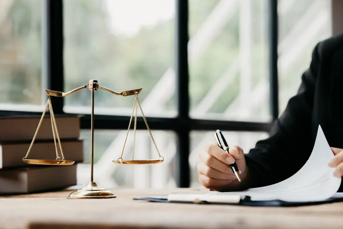 Balanza de la justicia sobre un escritorio junto a una persona que escribe en un documento, cerca de una ventana.