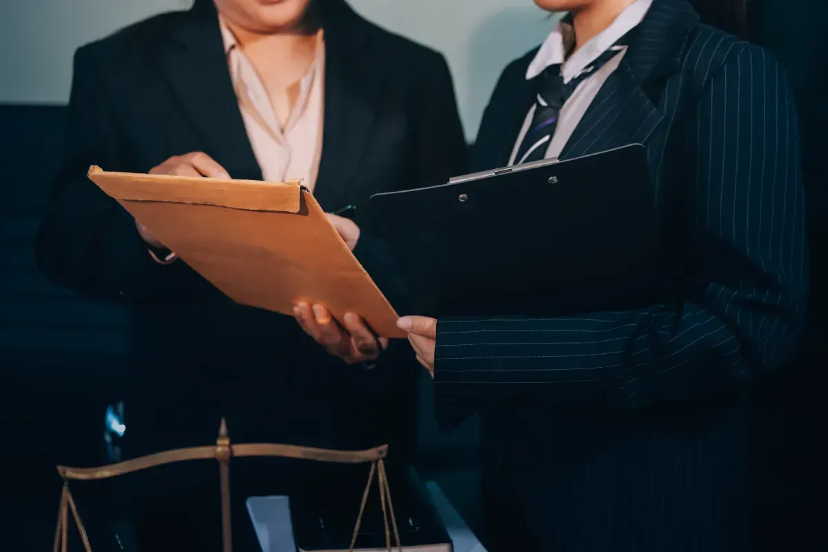 Dos personas con traje examinando documentos, cerca de la balanza de la justicia.