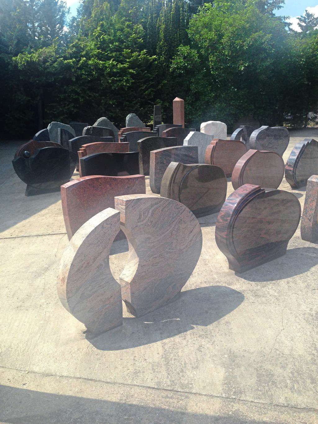 Eine Sammlung von Grabsteinen in verschiedenen Formen, die im Freien in den Farben Rot, Braun und Weiß vor einem Hintergrund aus Bäumen ausgestellt sind.