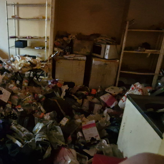 Pièce encombrée de déchets, de cartons et de sacs en papier éparpillés, avec des étagères le long des murs.