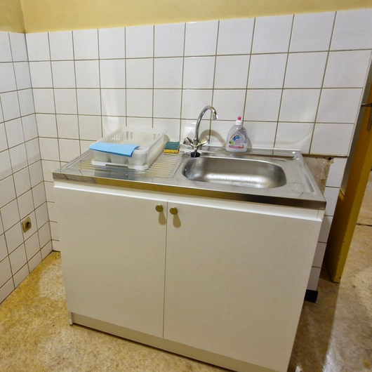 Petit évier de cuisine et meuble blanc sous un mur carrelé, avec un égouttoir à vaisselle et un savon sur le comptoir.