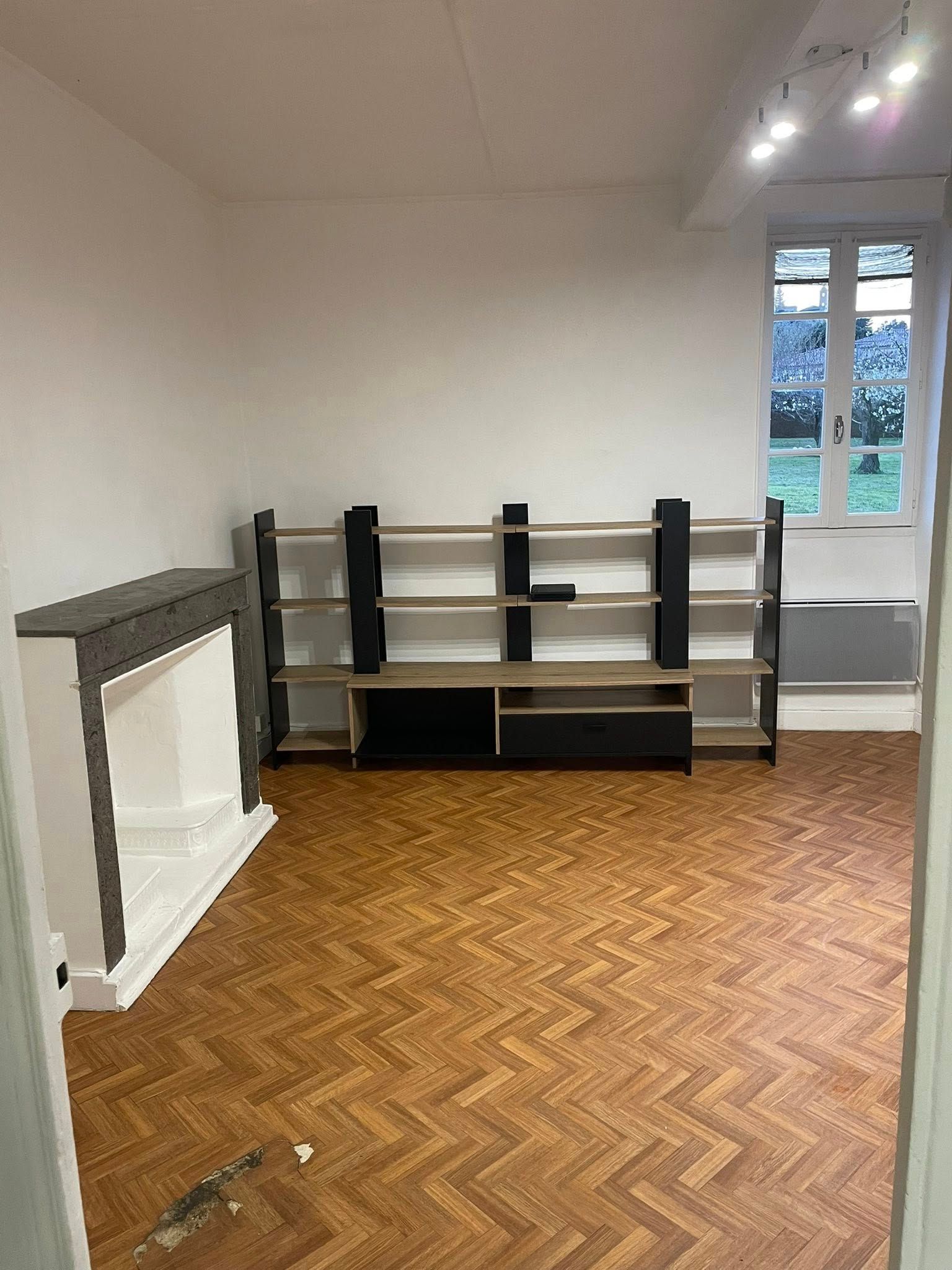 Pièce vide avec parquet à chevrons, des étagères noires et une petite fenêtre sur la droite.