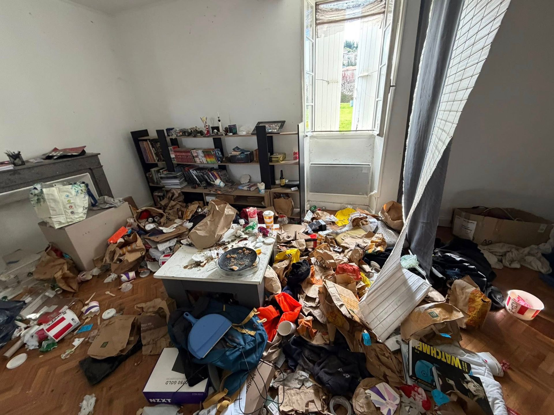 Pièce encombrée de déchets et d'objets éparpillés sur le sol, près d'une fenêtre aux stores ouverts.