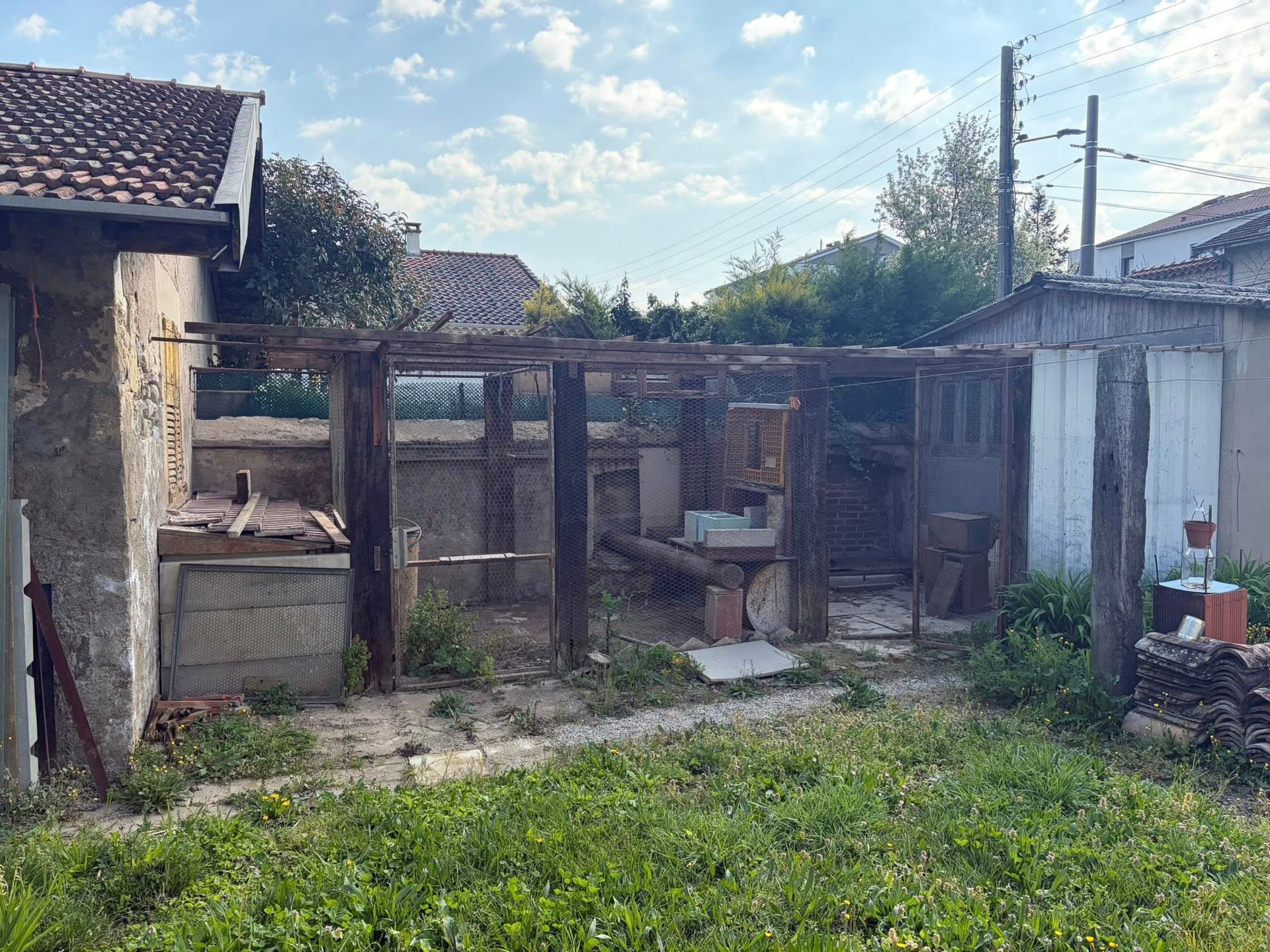 Des enclos à animaux en bois délabrés jouxtent une maison rustique, avec de l'herbe devant et un ciel partiellement nuageux.
