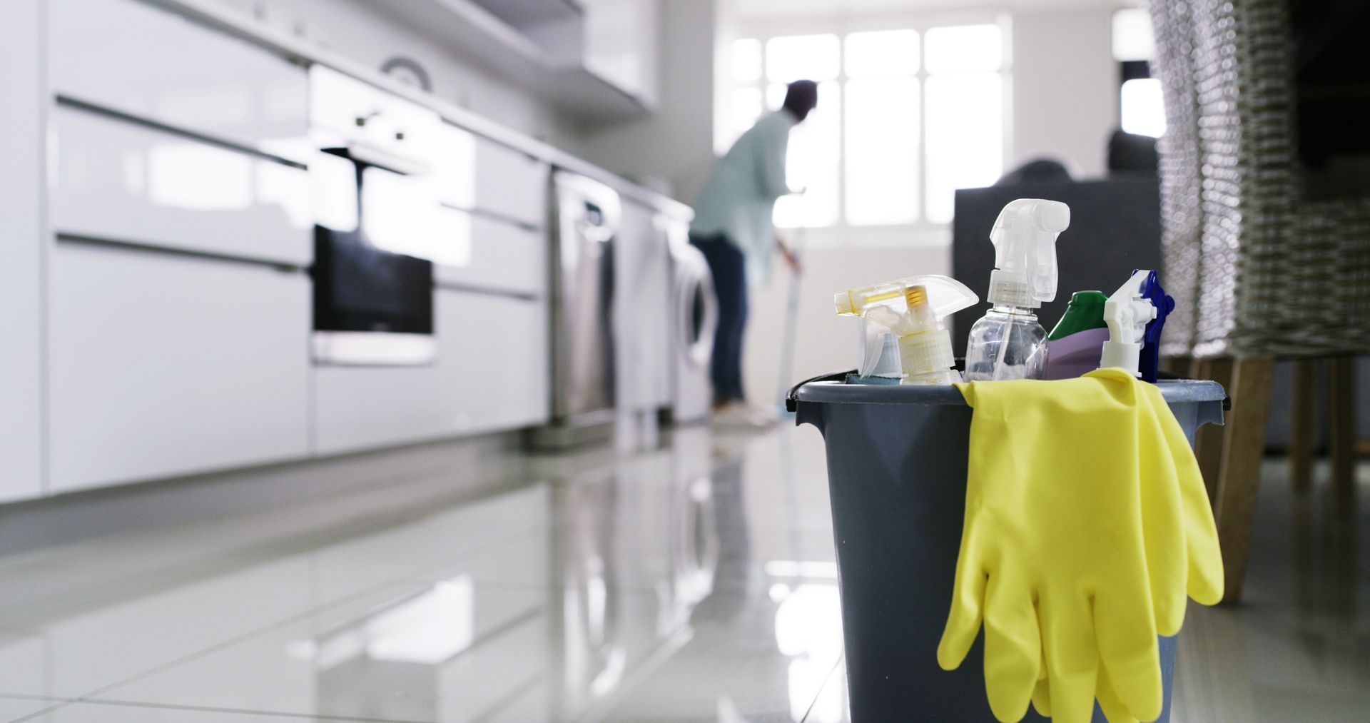 Des produits de nettoyage dans une cuisine, avec des gants jaunes accrochés à un seau et une personne qui range en arrière-plan.