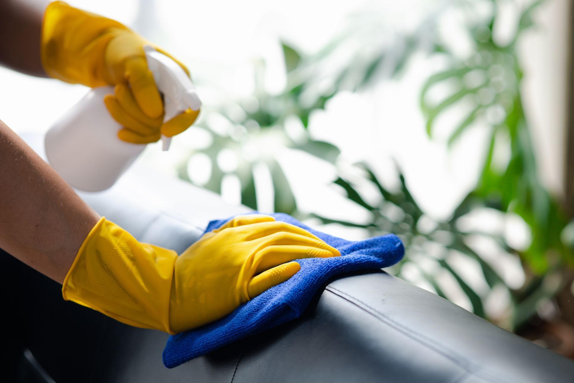 Un agent d'entretien avec des gants de ménage jaunes nettoie un canapé en cuir noir avec un chiffon bleu et un vaporisateur.
