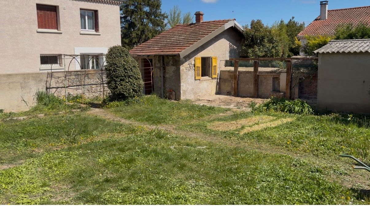 Petit jardin gazonné avec une dépendance en briques, un portail et une maison baignée de soleil.