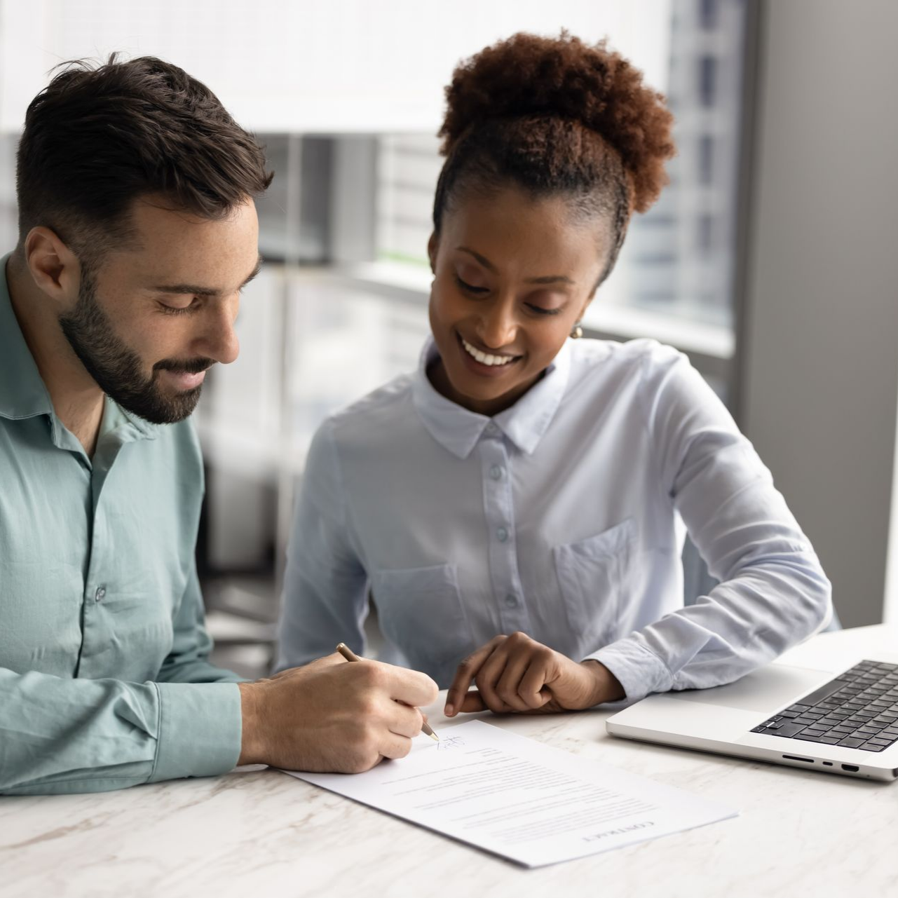 Un homme et une femme sourient, consultant ensemble un document à un bureau avec un ordinateur portable.