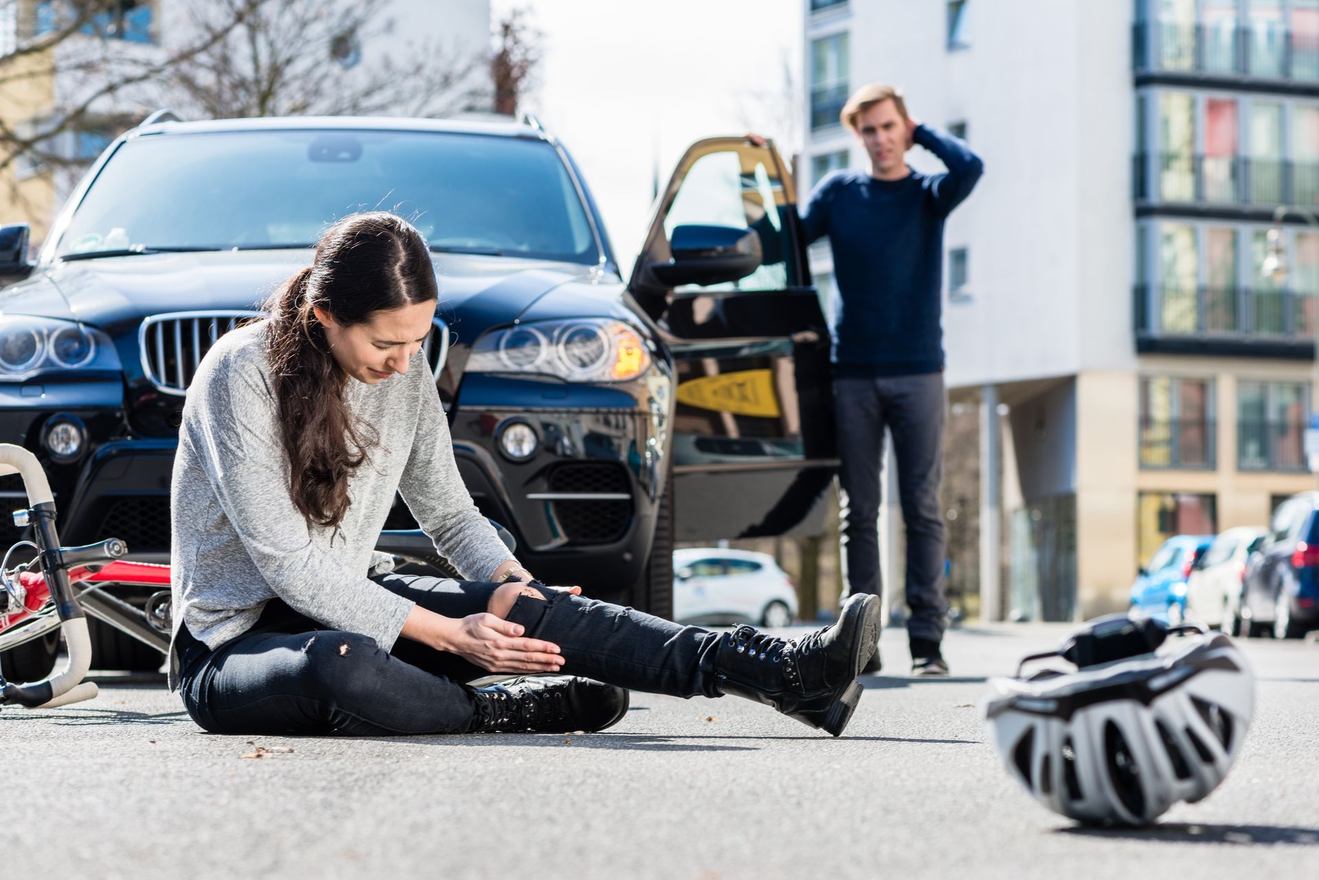 Une femme blessée, assise sur la route après une collision de voitures, son casque à proximité, un homme la regarde.