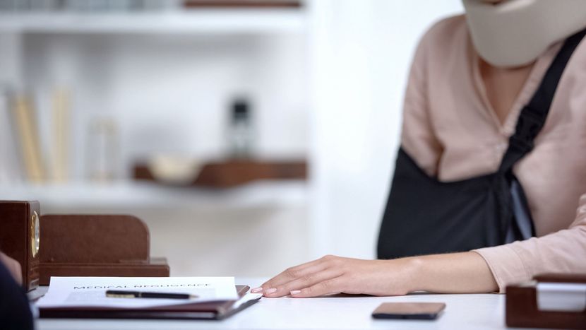 Une personne portant une minerve et une écharpe au bras est assise à un bureau avec des papiers et un téléphone.