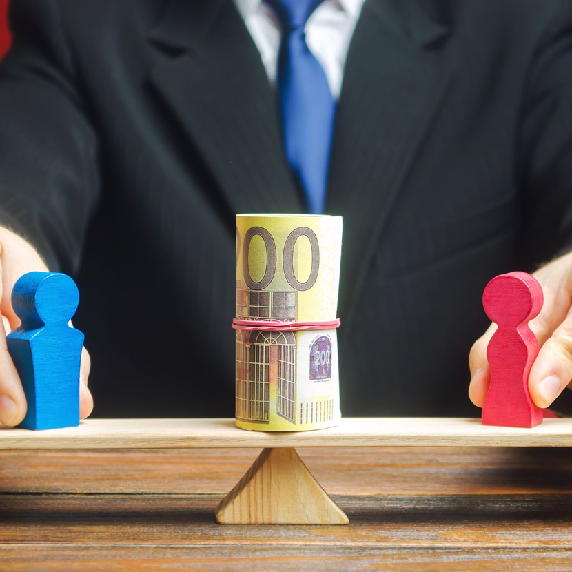 Un homme d'affaires tient une liasse de billets en équilibre entre des figurines en bois bleues et rouges sur une balance.