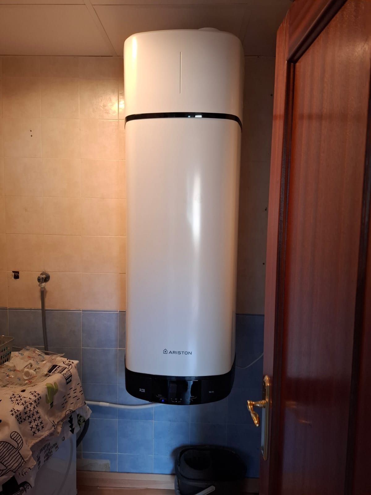 Calentador de agua blanco montado en una pared de azulejos beige y azules junto a una puerta marrón.