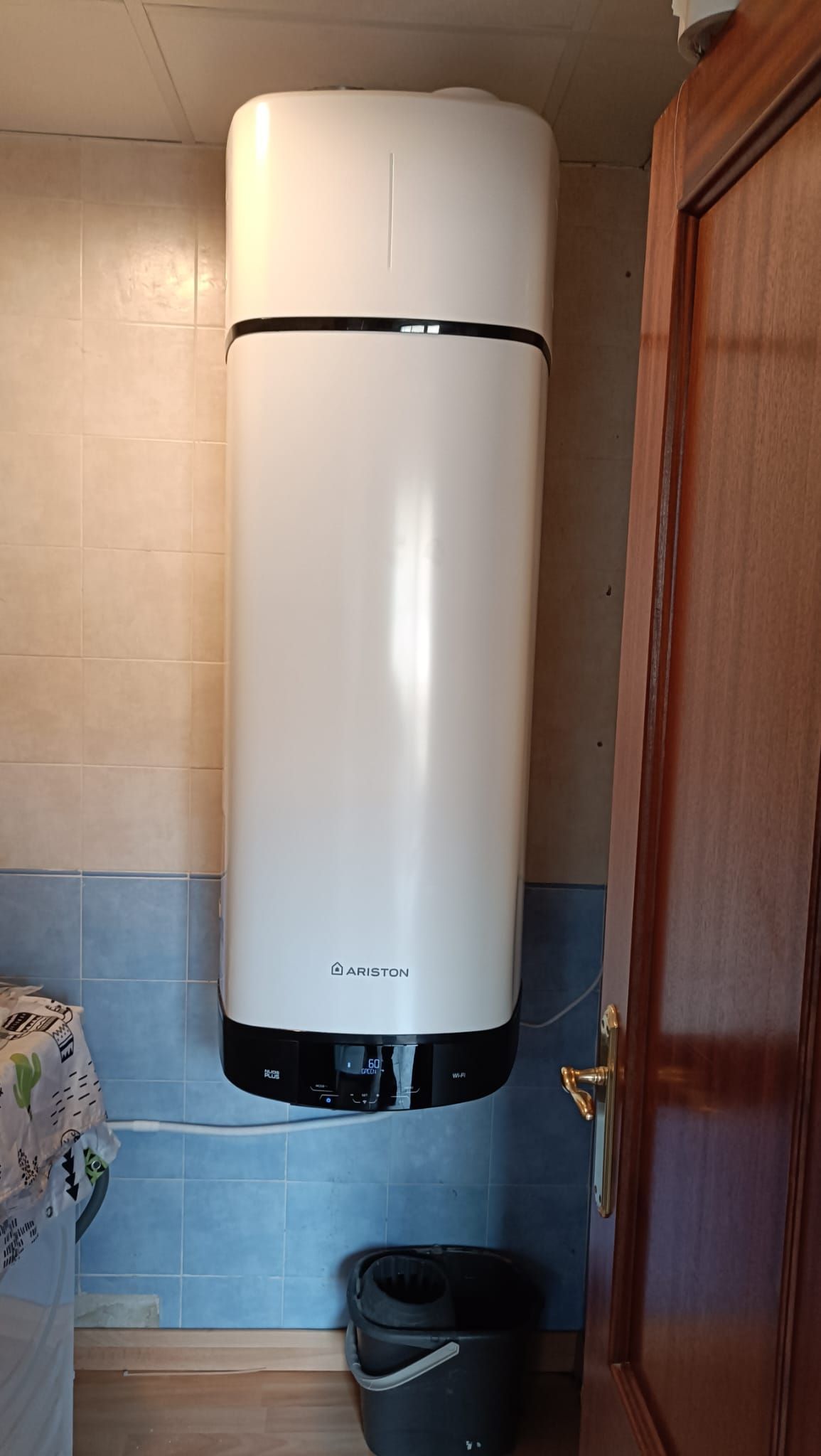 Calentador de agua blanco montado en una pared de azulejos en una habitación, con una puerta parcialmente visible y un balde debajo.