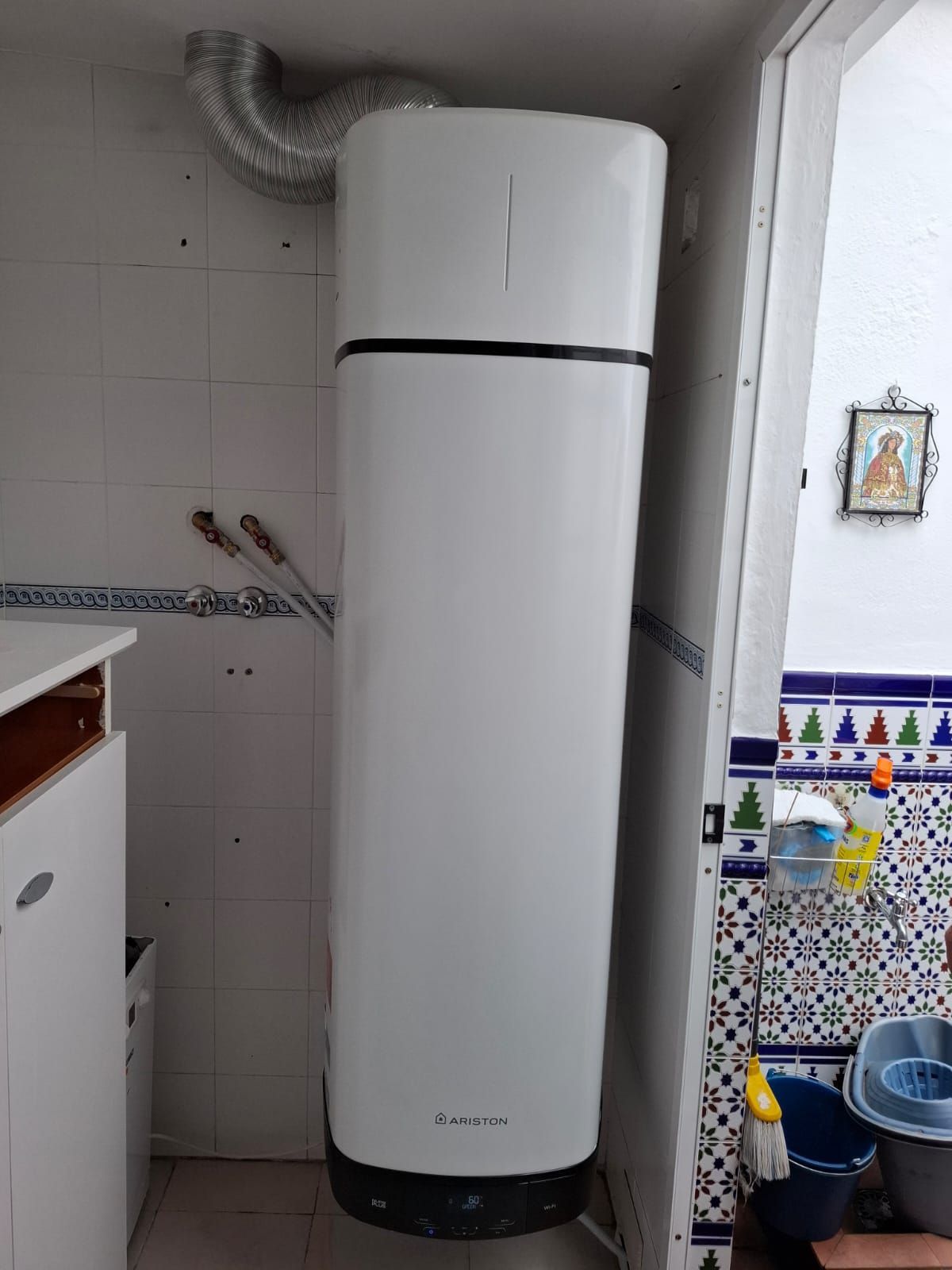 Calentador de agua alto y blanco con tubo de escape, sobre pared de azulejos al lado de una puerta abierta.