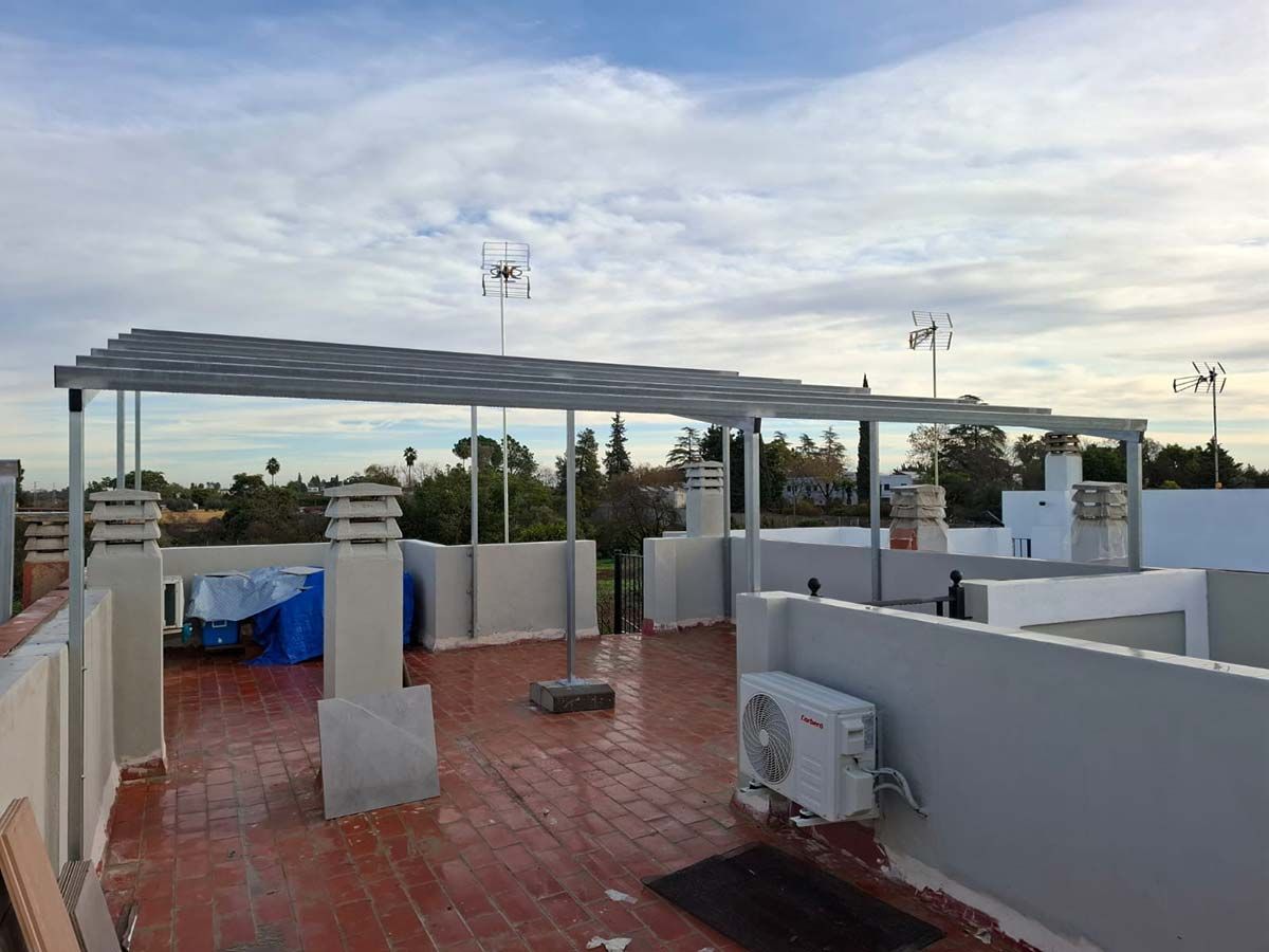 Azotea con pérgola metálica, equipo de aire acondicionado y varias chimeneas bajo un cielo nublado.