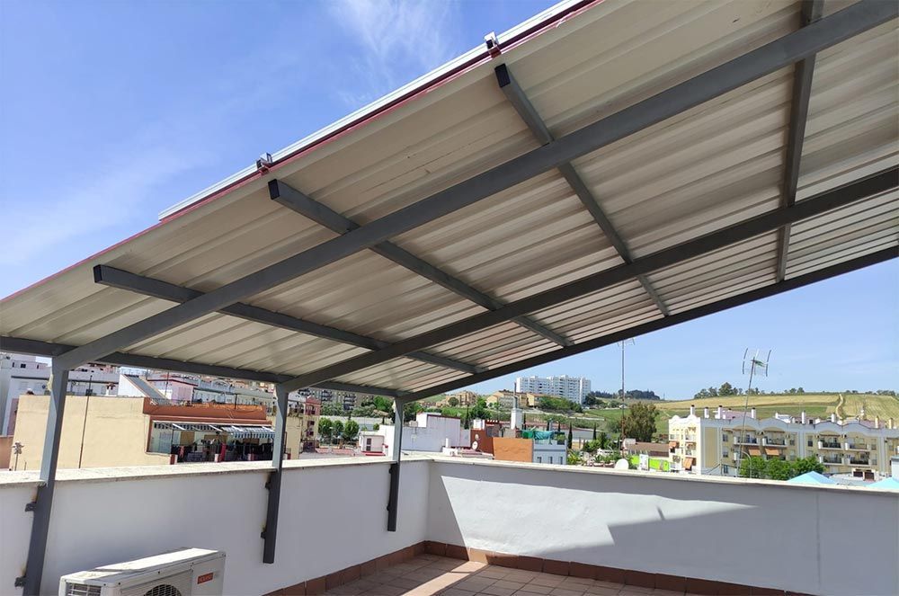 Una estructura de techo de metal y cartón corrugado proporciona sombra a un patio, con un cielo brillante de fondo.