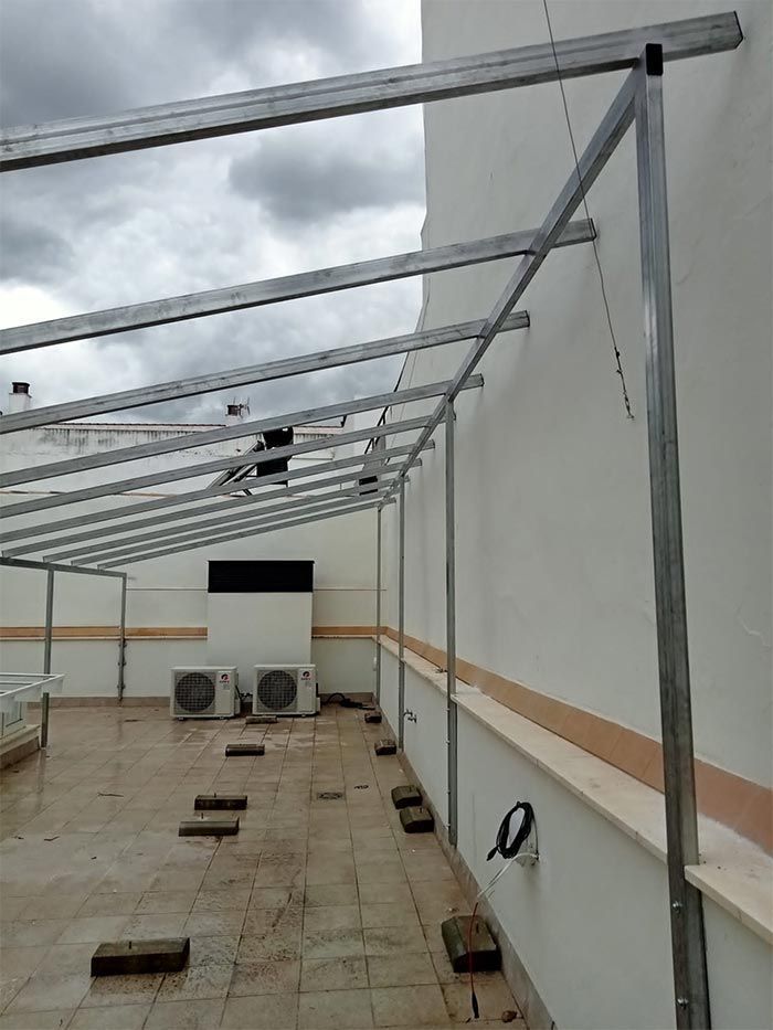 Estructura metálica fijada al exterior de un edificio blanco. Marquesina parcialmente construida sobre un patio de baldosas con aire acondicionado.