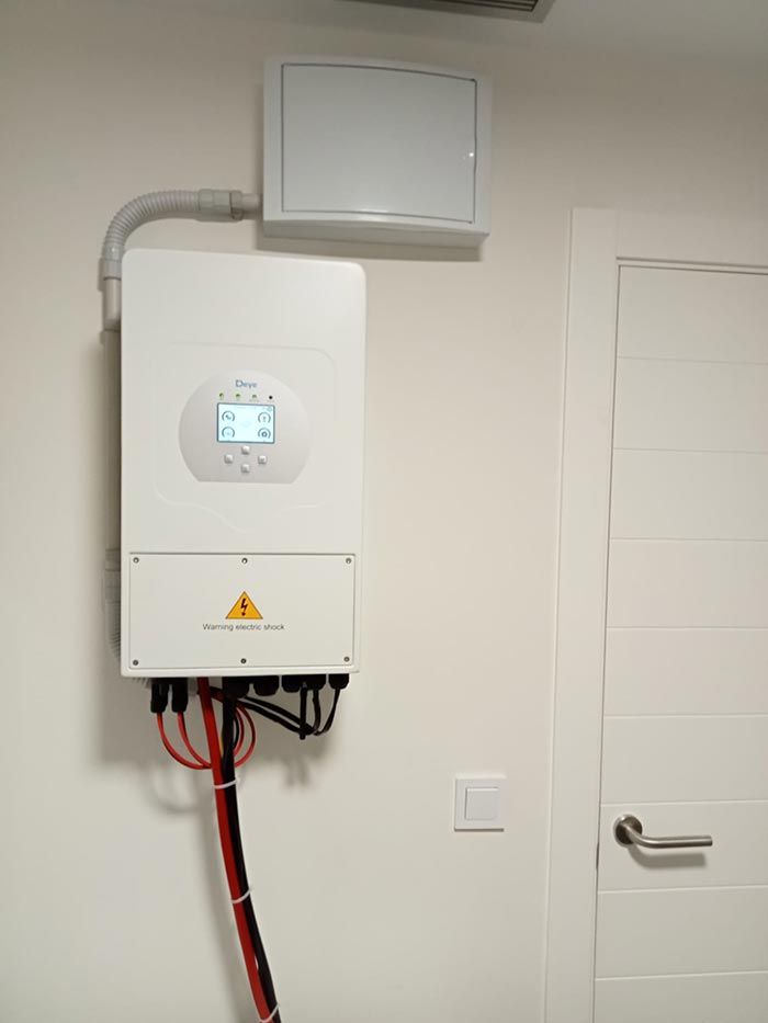 Inversor solar blanco montado en la pared, con cables conectados y una pequeña pantalla. Junto a él hay un panel eléctrico y una puerta blanca.