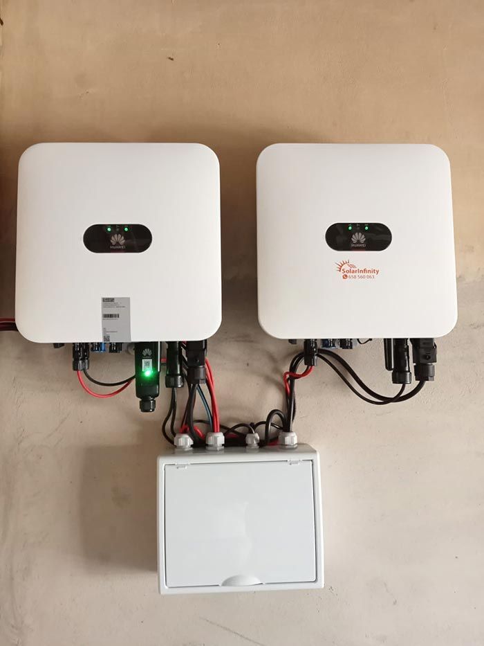 Dos inversores solares blancos y una caja de conexiones montados en una pared beige, conectados mediante cables.