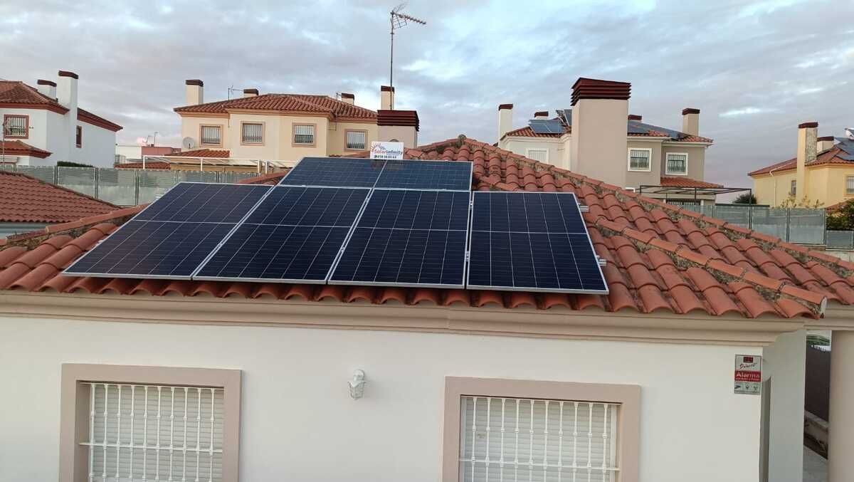 Paneles solares sobre un techo de tejas rojas de una casa blanca, rodeada de otras casas bajo un cielo nublado.