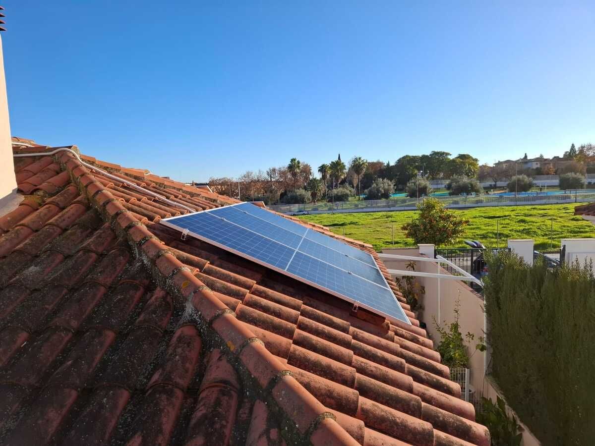 Paneles solares sobre un techo de tejas rojas con un cielo azul brillante y un campo verde al fondo.