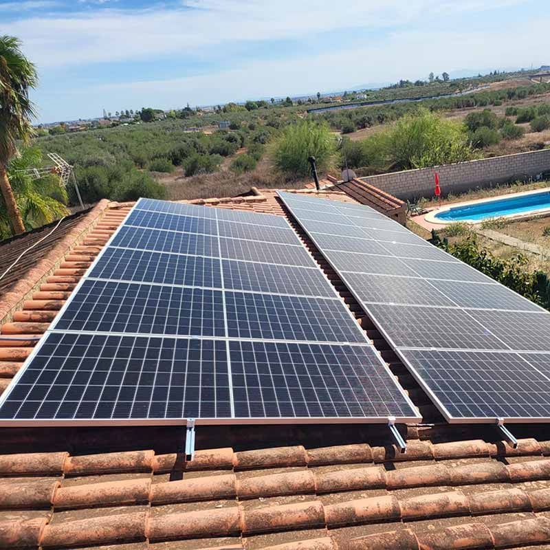 Paneles solares instalados sobre un tejado de tejas rojas, con una piscina y un campo al fondo.